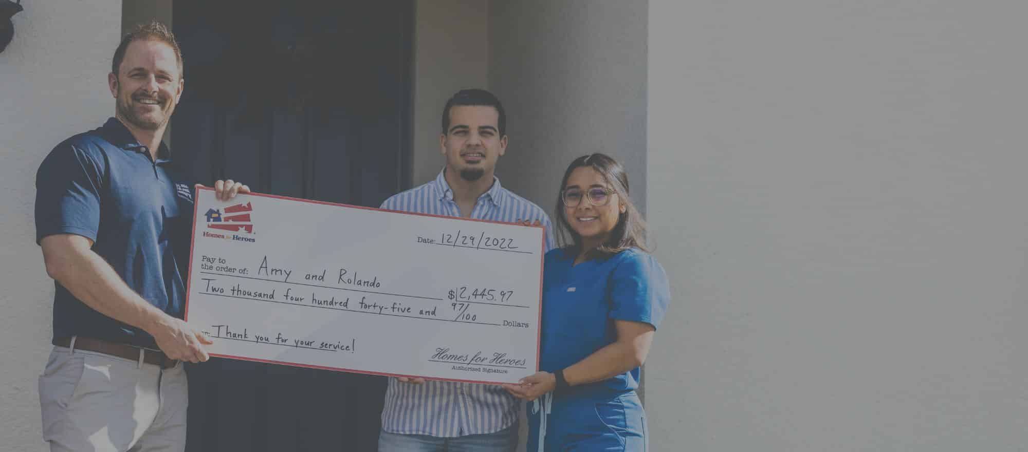 Homes for Heroes male real estate agent holding big check with hero home buyer couple in front of new home