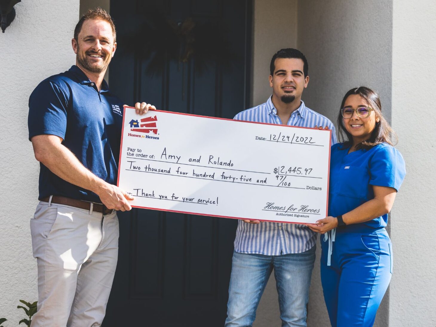 Amy and Rolando holding big check outside new home with Homes for Heroes specialist  