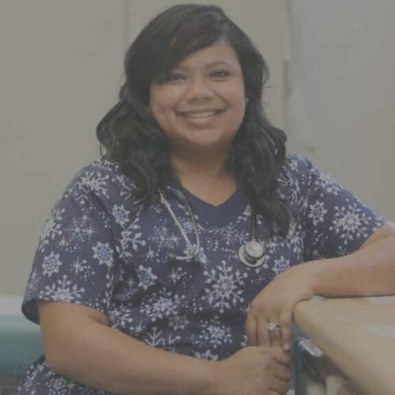 woman school nurse in student care area smiling leaning on table purple patterned scrubs