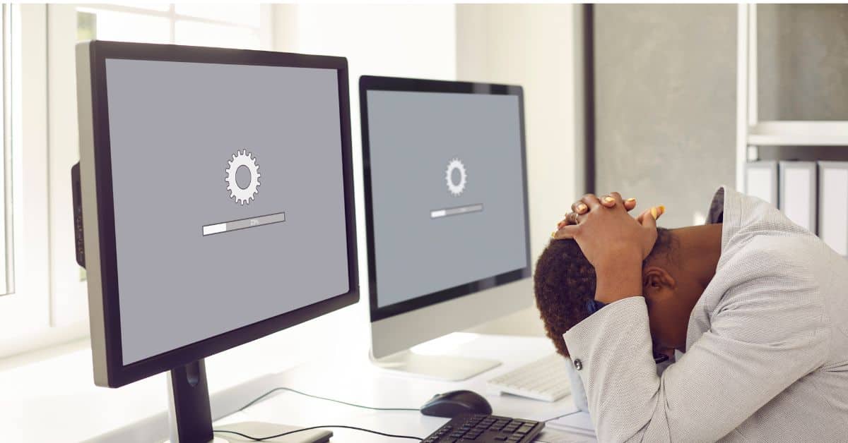 person sitting with head on desk frustrated by slow load site speed shown on two computer monitors in office