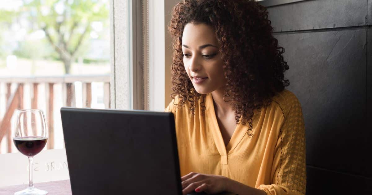 excited woman sitting in restaurant with glass of wine working on laptop signing up to receive free lead magnet asset from real estate agent