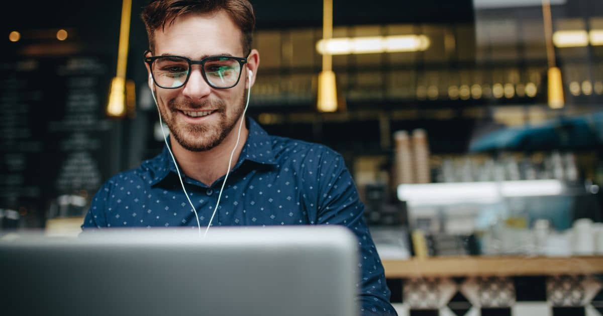 male real estate agent smiling wearing buds in ears in coffee shop on laptop looking at websites Homes for Heroes